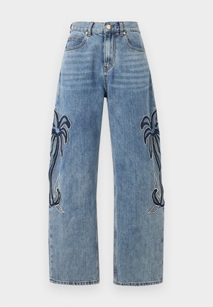 Nő áll, visel egy ujjatlan tengerészkék kötött felsőt és bőszárú kék farmert, melyen nagy sötét pálmamutatók vannak a lábszárakon.