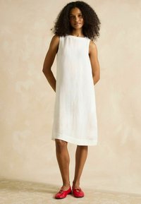 Weisses Leinenkleid mit ärmellosem Design, gerader Form und einem Saum, der unter dem Knie endet. Kombiniert mit roten Ballerinas mit Schleifendetail.