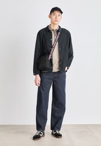 Camicia a bottoni nera, polo beige, jeans in denim blu larghi, borsa a tracolla nera con tracolla logo, scarpe da ginnastica bianche e nere, cappellino.