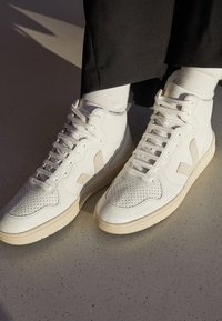 Vita högblanka sneakers med beige sulor och sidodetaljer, bärs med vita strumpor och svarta byxor, stående på en ljus prickig golv.