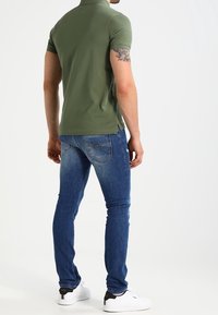 Oliwkowa koszulka polo z krótkim rękawem, zestawiona z niebieskimi obcisłymi dżinsami i białymi trampkami z czarnymi akcentami. Widoczny tatuaż na lewym ramieniu.