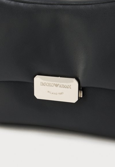 Μαύρη δερμάτινη τσάντα χειρός με πλακέτα λογότυπου από μεταλλικό υλικό σε ασημί απόχρωση, με ανάγλυφο κείμενο. Ομαλή υφή και στρογγυλεμένο σχήμα.