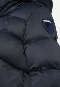 Chaqueta acolchada azul marino con secciones acolchadas, que presenta una etiqueta de tela y un parche bordado con el logo en el hombro izquierdo. Textura suave.