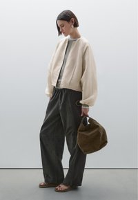 Vrouw in een losse beige jas, brede donkere broek, sandalen, die een grote bruine suède tas vasthoudt, staand tegen een eenvoudige lichtgrijze muur.