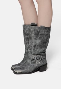Botas de cuero negro desgastadas y rasgadas hasta la mitad de la pantorrilla con punteras de metal plateado, detalles de correas y remaches, y tacones cuadrados sobre piernas descubiertas.