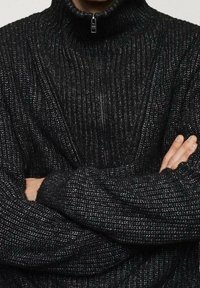 Schwarzer gestrickter Pullover mit hohem Kragen, vertikalen Rippen, einem halben Reißverschluss und einem strukturierten Muster.