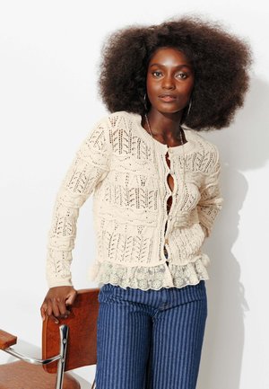 Femme aux cheveux bouclés et volumineux portant un gilet en maille crème sur une blouse en dentelle et un pantalon bleu à rayures, appuyée sur une chaise en bois.