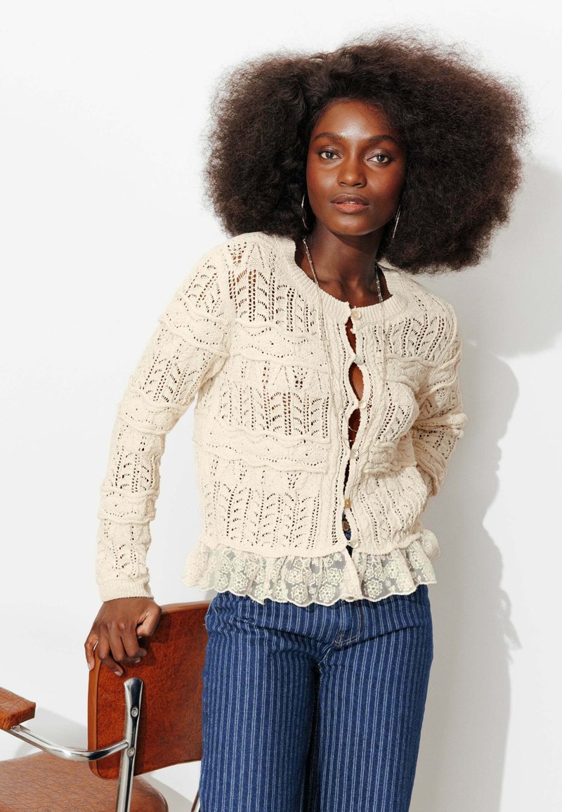 Femme aux cheveux bouclés et volumineux portant un gilet en maille crème sur une blouse en dentelle et un pantalon bleu à rayures, appuyée sur une chaise en bois.