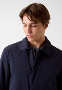 Marineblauwe lichte jas met een opstaande kraag, ritssluiting en subtiele textuur. Heeft een gestroomlijnd ontwerp zonder zichtbare patronen.