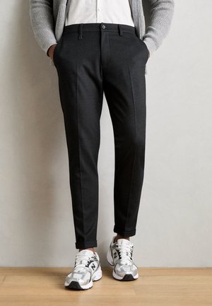 Uomo che indossa pantaloni neri su misura, camicia bianca, cardigan grigio e sneakers bianche New Balance, in piedi su pavimento di legno.