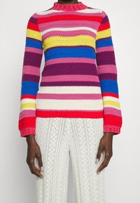 Flerfärgad stickad tröja med vågiga ränder i röd, rosa, gul, blå och lila. Har en hög krage och texturerade ärmar.