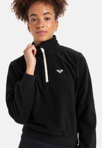Black fleece pullover with a high collar, quarter zip closure, and white accent. Features ribbed cuffs and a small logo on the chest.