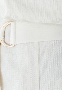 Witte getextureerde stof met een golvend patroon, voorzien van een effen witte tailleband en een ronde metalen gesp in roségouden afwerking.