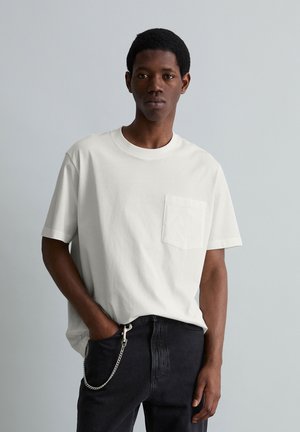 Junger Mann trägt ein schlichtes weißes T-Shirt mit Tasche und schwarze Jeans mit einer silbernen Kette, steht vor einem neutralen hellgrauen Hintergrund.