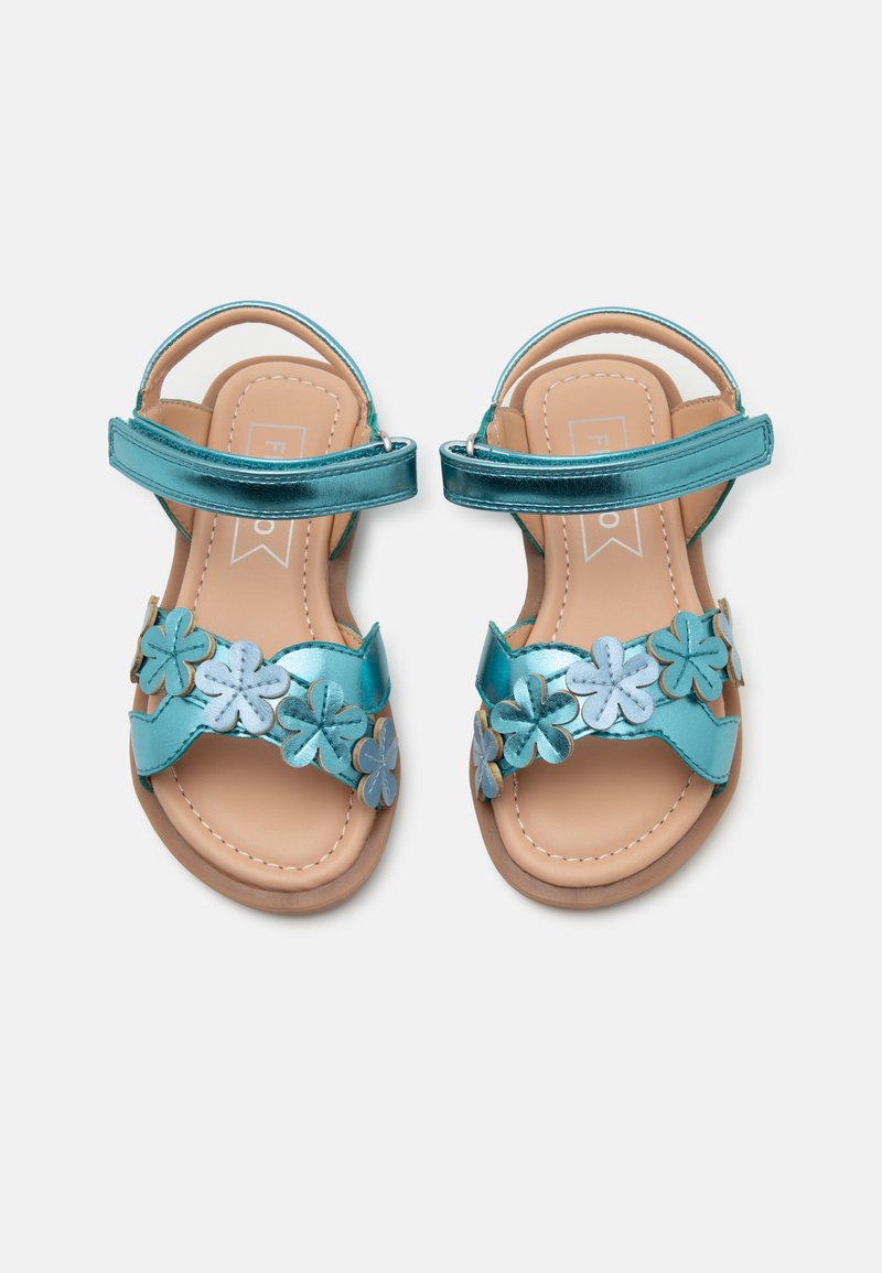 Turkise sandaler med metallisk remmer, blomsterdekorasjoner og polstret såle. Har justerbare ankelremmer og flat såle.