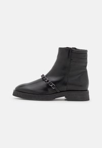 Bota de piel negra hasta el tobillo con tacón bajo, cremallera lateral y cadena decorativa en la parte superior, con la marca Marc O'Polo en el collar.