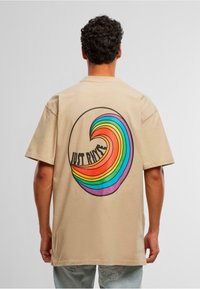 Beige bomull T-skjorte med et stort, flerfarget bølgedesign på baksiden, som inkluderer røde, oransje, gule, grønne og blå striper med tekst.