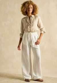 Svetlohnedá pruhovaná košeľa na gombíky s volánikovým detailom, v kombinácii s voľnými bielymi ľanovými nohavicami a hnedými sandálmi so vzorom.
