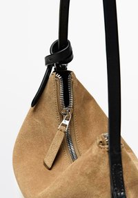 Tan mockaskinnsväska med svart läderrem, försedd med en silverfärgad dragkedja och en mockaskinnsdragkedjehållare. Designen är rundad och minimalistisk.