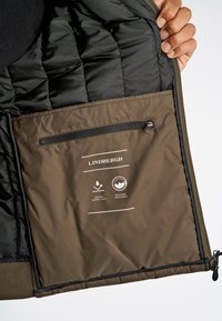 Fodrad jacka inuti med svart tyg och en brun ficka med dragkedja, skötselråd och en logotyp. Vattenavvisande material.