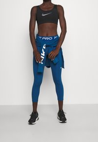 Svart sportbehå med silverlogga, blå leggings och en knuten blå sweatshirt. Svarta löparskor med vita detaljer. Slät, åtsittande textil.