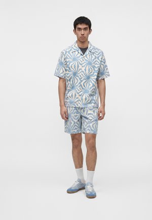 Ung mand står iført en blå- og hvidmønstret kortærmet skjorte og matchende shorts med hvide sokker og blå sneakers.