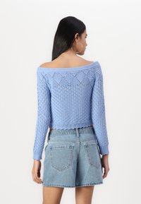 Blå stickad tröja med off-shoulder-design och strukturerat diamantmönster, långa ärmar och kort längd. Matchas med ljusa denimm shorts.