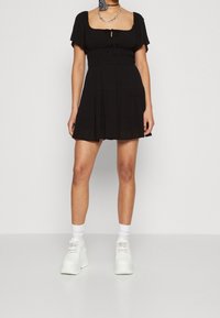 Kvinna som bär en kort svart klänning med puffärmar, vita strumpor och vita plattformssneakers, stående framför en neutral bakgrund.