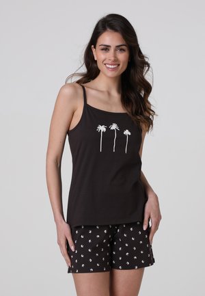 Donna sorridente con capelli lunghi e scuri, che indossa una canotta nera con tre palme bianche e pantaloncini neri con piccoli motivi di palme bianche.