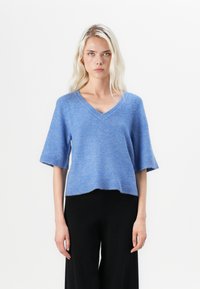Light blue knitted sweater with a V-neck, short sleeves, and a relaxed fit. Features a ribbed hem and smooth texture. Paired with black trousers.