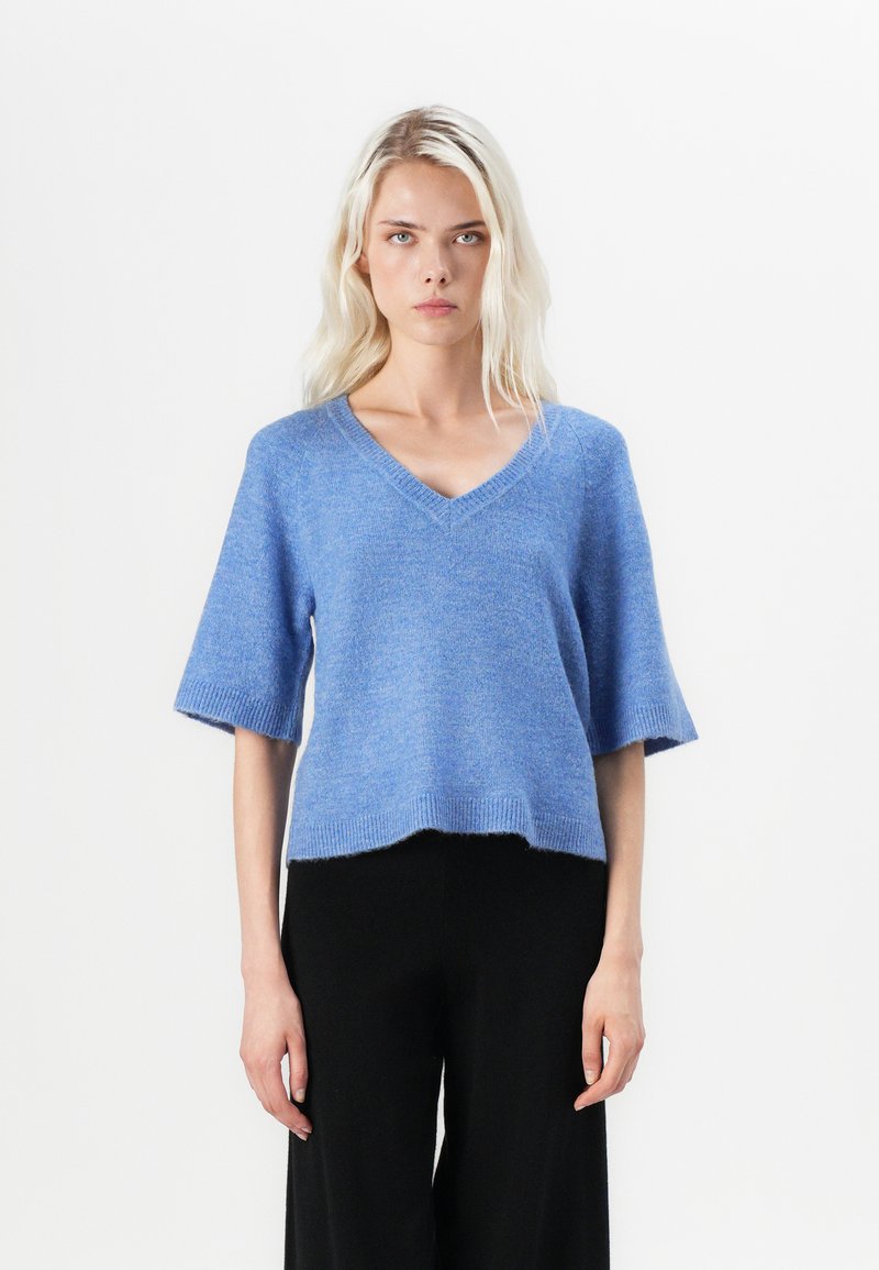 Light blue knitted sweater with a V-neck, short sleeves, and a relaxed fit. Features a ribbed hem and smooth texture. Paired with black trousers.
