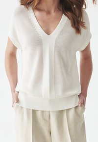 Žena v volném krátkém bílém pleteném tričku s hlubokým výstřihem do V a béžových lněných kalhotách, ruce v kapsách.