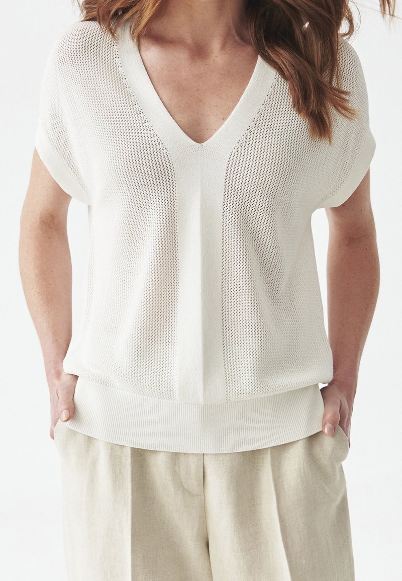 Žena v volném krátkém bílém pleteném tričku s hlubokým výstřihem do V a béžových lněných kalhotách, ruce v kapsách.