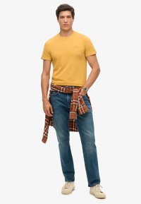 Camiseta amarilla de cuello redondo, vaqueros azules y zapatillas crema. Camisa roja de cuadros atada a la cintura. Estilo casual clásico.