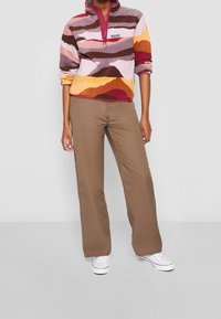 Persona con un pulóver de forro polar de patrón multicolor, pantalones marrones y zapatillas blancas, de pie contra un fondo claro y liso.