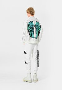 Sudadera blanca con un gran diseño de alas de lentejuelas negras y turquesas en la parte de atrás, combinada con pantalones joggers blancos que presentan estampados de alas negras y rotos.