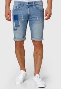 Pantaloni scurți de blugi bleu deschis cu accente uzate și un design patchwork pe coapsa stângă. Margini rulate și stil cu cinci buzunare. Adidași albi.