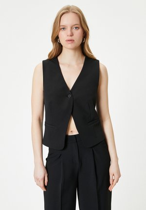 Zwarte mouwloze vest met een V-hals, enkele knoopsluiting en een spitse onderkant, voorzien van twee voorzakken en een gladde stofstructuur.