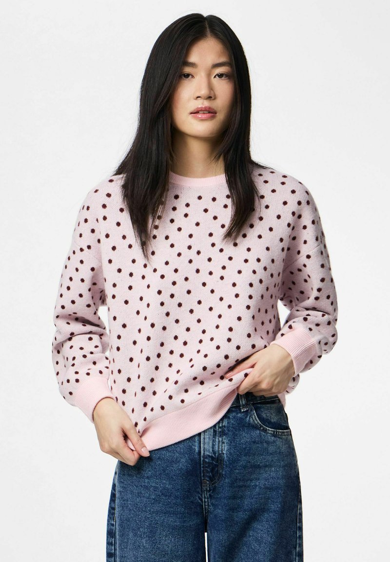 Pull rose clair avec un col rond, décoré de pois marron foncé, poignets et ourlet côtelés. Porté avec un jean en denim bleu.
