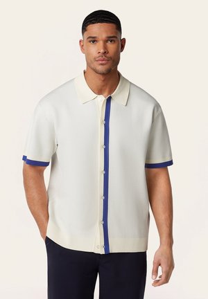 Man die een wit overhemd met korte mouwen draagt met blauwe bies op de knoopsluiting en zoom van de mouwen, gecombineerd met een donker broek, staand tegen een effen achtergrond.