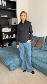 Kobieta o blond włosach, ubrana w czarną koszulę na guziki i szerokie niebieskie dżinsy, stoi w salonie z turkusową sofą i regałem na książki.