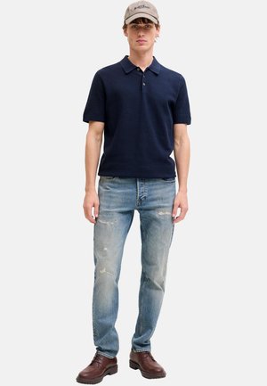 Jonge man die een marineblauw polo met korte mouwen draagt, lichte distressed jeans, bruine schoenen en een beige pet, kijkend naar voren staand tegen een witte achtergrond.