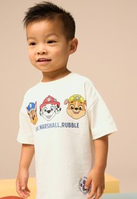 White cotton t-shirt featuring cartoon dog designs in color. Text reads "Chase, Marshall, Rubble" in blue. Short sleeves and round neckline.