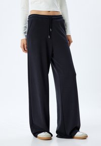 Wijdvallende sweatpants in donkerblauwe stof; voorzien van een elastische tailleband met trekkoord en zijzakken, gedragen met witte lage sneakers.
