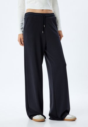 Wijdvallende sweatpants in donkerblauwe stof; voorzien van een elastische tailleband met trekkoord en zijzakken, gedragen met witte lage sneakers.