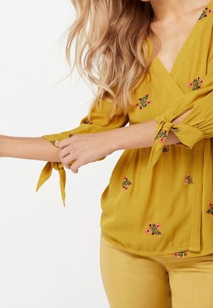 Vrouw die de mouw van een mosterdgele blouse met bloemenborduursel en gestrikte strikjes aanpast, gecombineerd met een bijpassende gele broek.