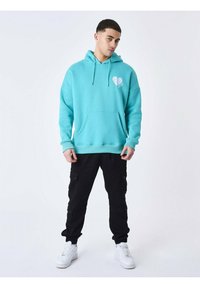 Sudadera con capucha turquesa, con un bolsillo delantero y un gráfico de corazón blanco, combinada con pantalones cargo negros y zapatillas blancas.
