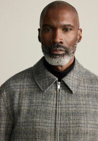 Gray plaid jacket with a collared neckline and front zipper. Textured fabric with a subtle pattern, worn over a black turtleneck.