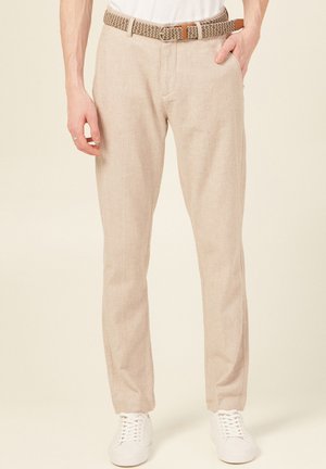 Pantaloni in lino beige a vestibilità rilassata, con tasche laterali, una fascia in vita con cintura intrecciata, abbinati a sneakers bianche.