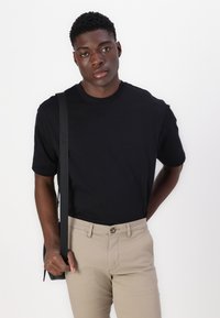 Camiseta de algodón negra con mangas cortas y cuello redondo, combinada con pantalones beiges. Modelo sosteniendo un bolso con una correa negra. Fondo liso.